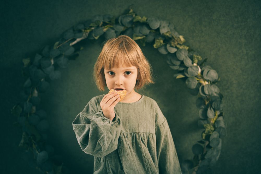 Child in gray dress standing under floral arch against dark background.