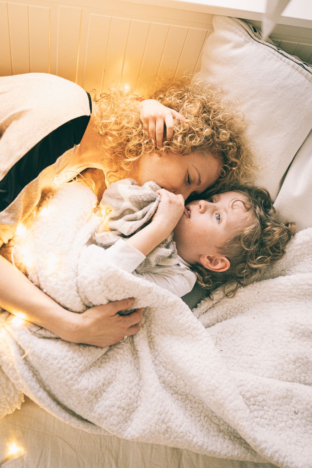 Two figures rest peacefully on white bedding illuminated by warm fairy lights.