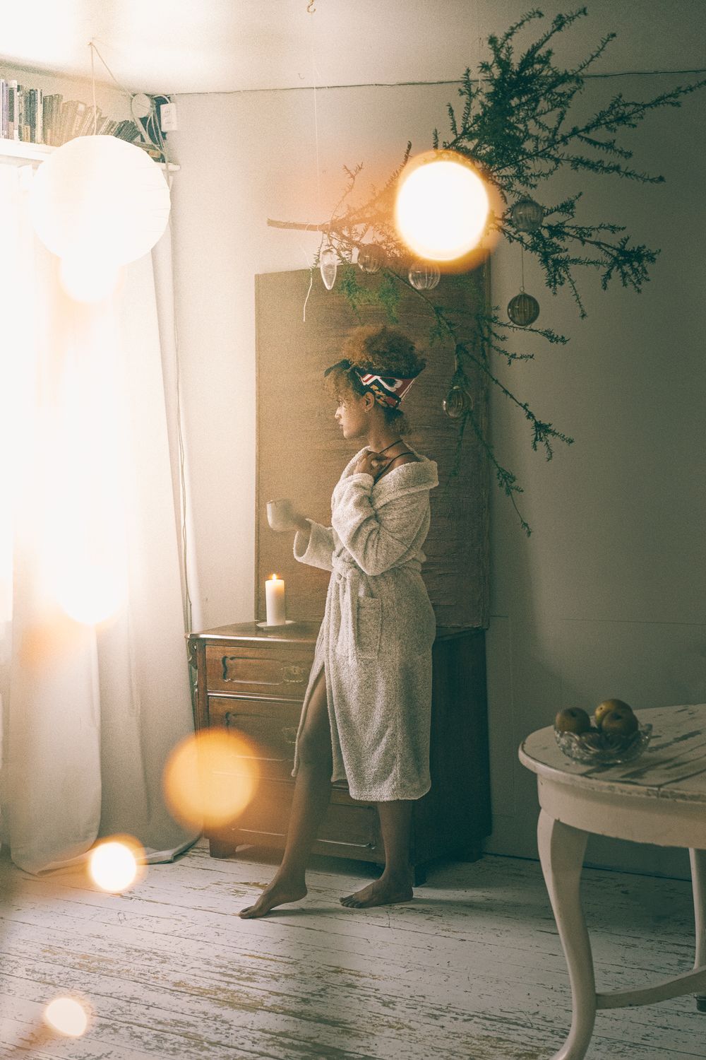 Person in bathrobe standing by window with sunlight and decorative branches.