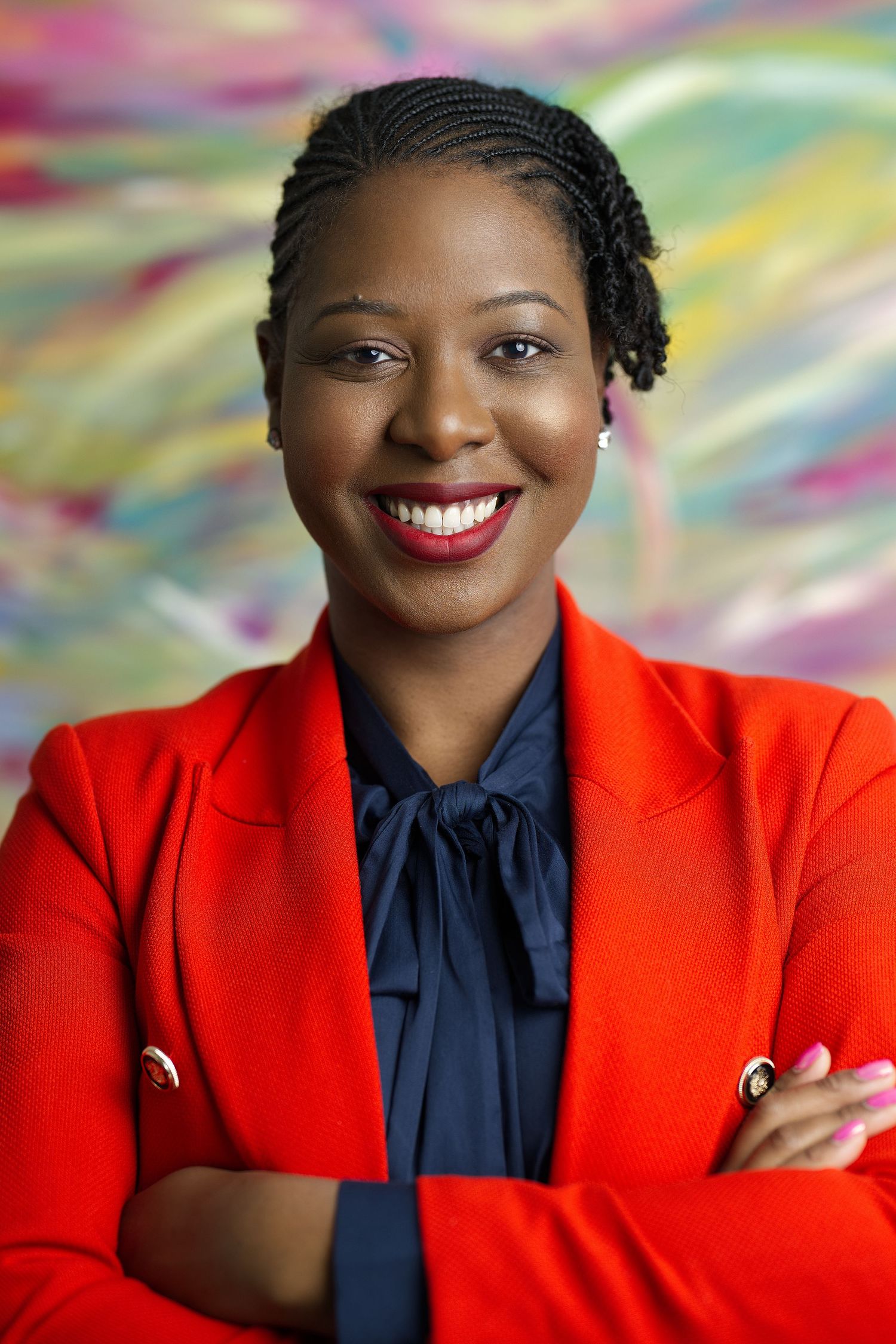 Professional portrait against colorful background showing bright red blazer and navy blouse with confident crossed arms pose.