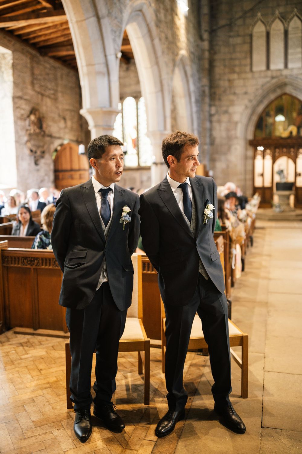 Two groomsmen in matching dark suits stand at the altar of a historic stone church with gothic arches.
