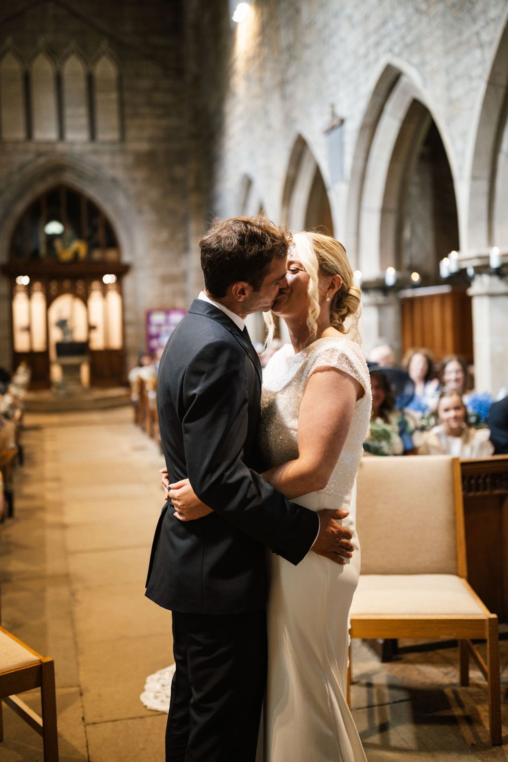 A couple shares their first kiss during their wedding ceremony in a historic stone church with arched columns.