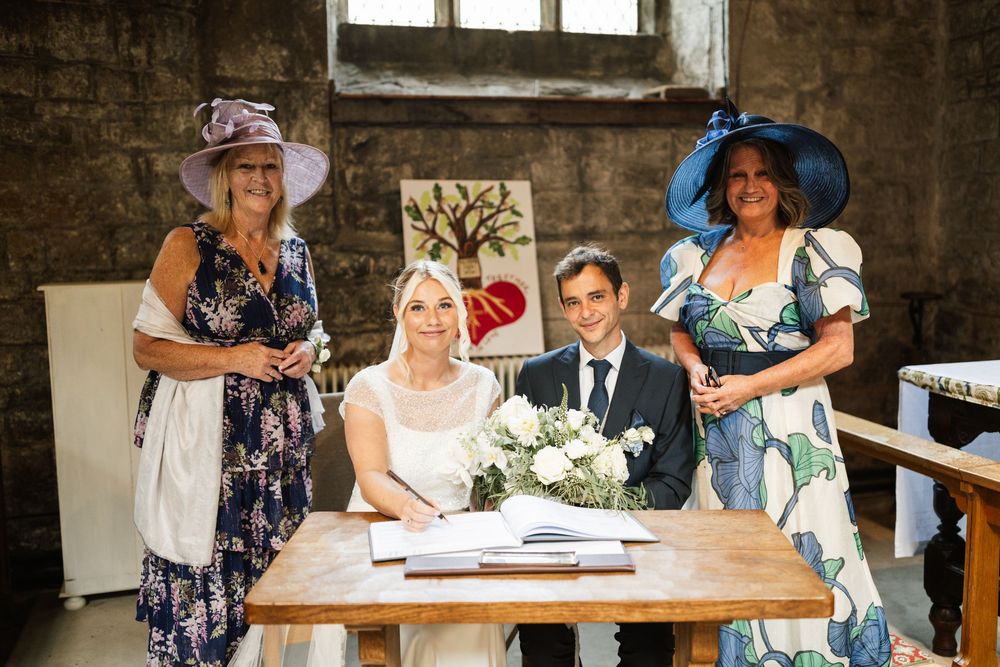 Wedding party gathered around a wooden table during marriage document signing in a rustic brick interior.