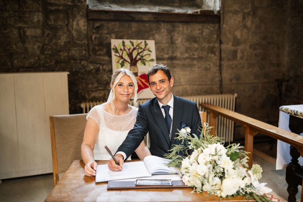 Couple signing marriage documents at a rustic venue with white floral arrangements on the table.