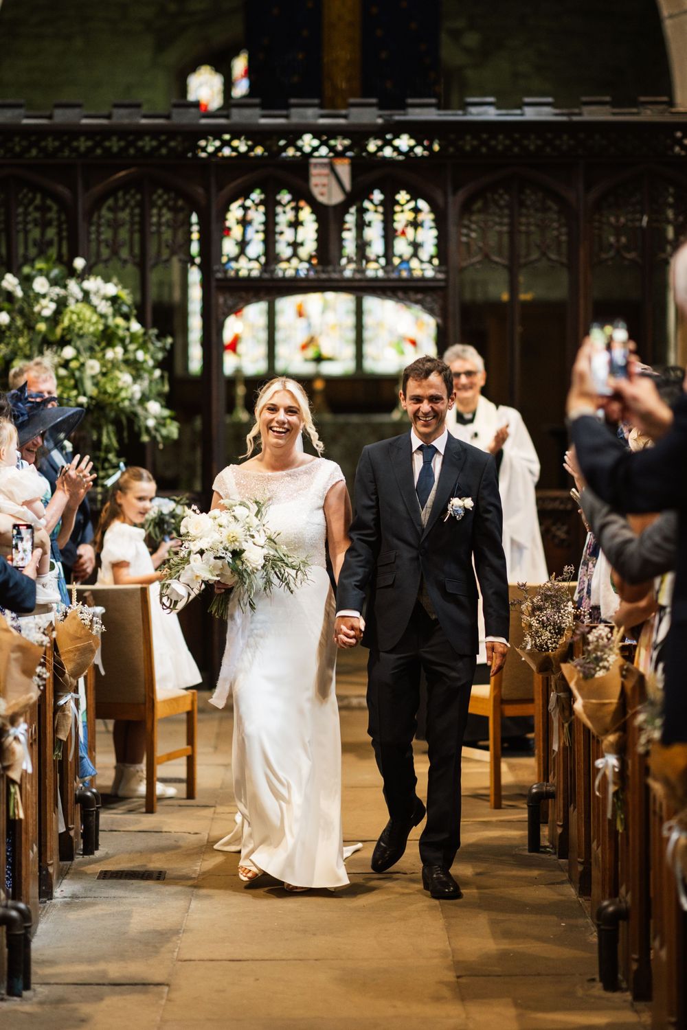 A wedding couple walks down the church aisle as guests look on beneath stained glass windows and ornate woodwork.
