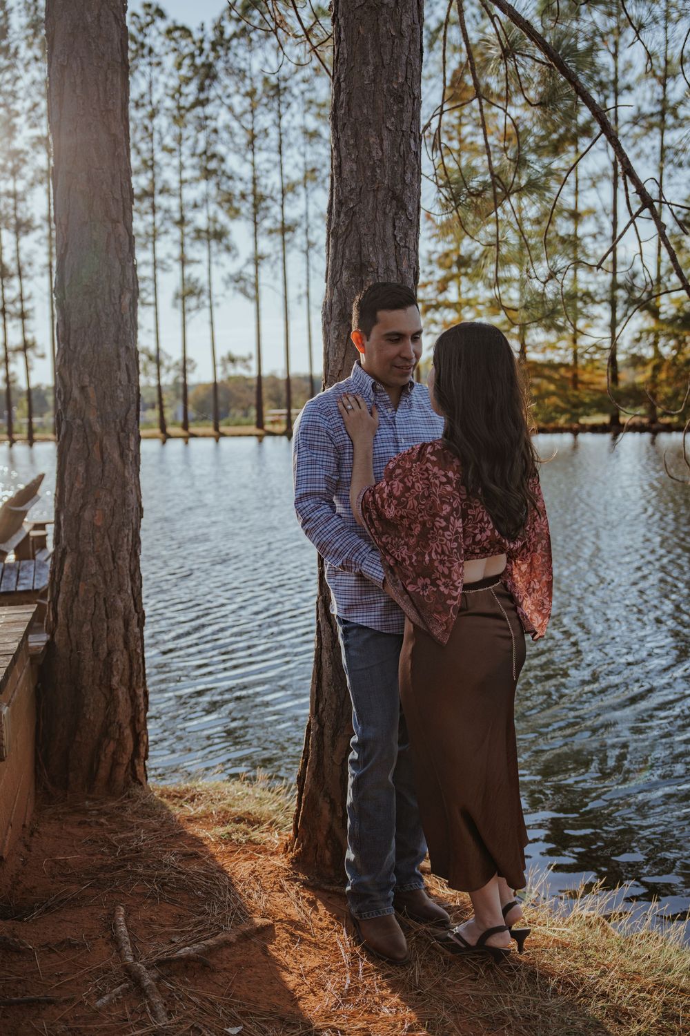 A couple shares an intimate moment by a lakeside with tall pine trees creating a natural backdrop.