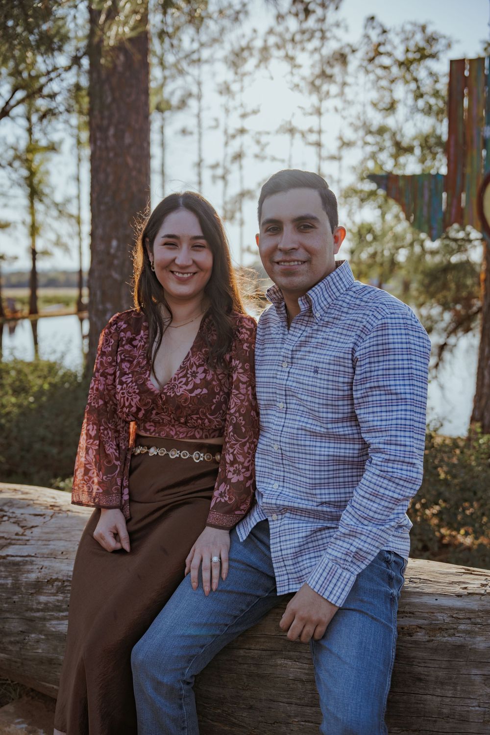 A couple sits together on a fallen log with pine trees and a lake in the background.