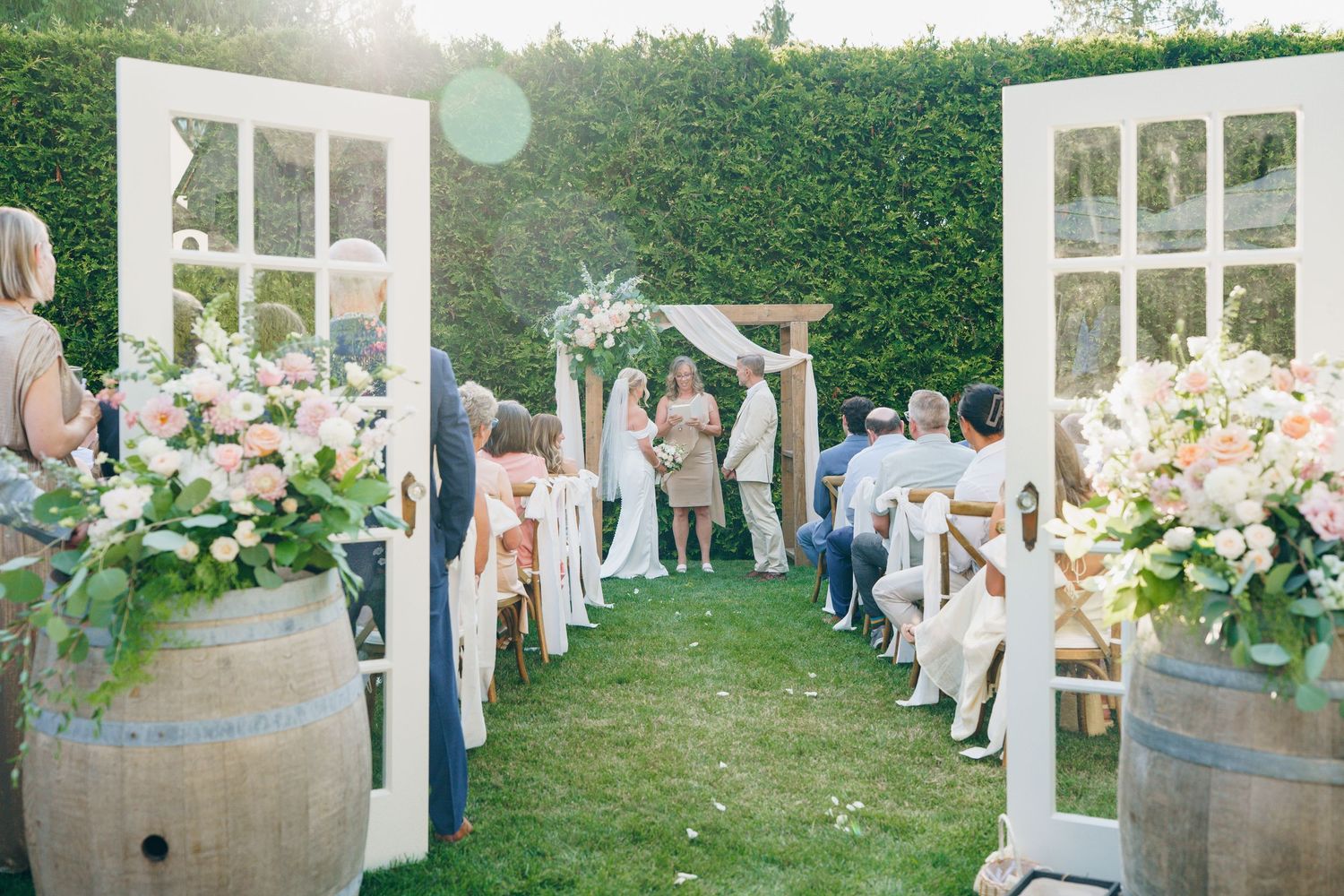 Wedding ceremony setup in a garden with white chairs, floral arrangements, and sunlight filtering through the trees.