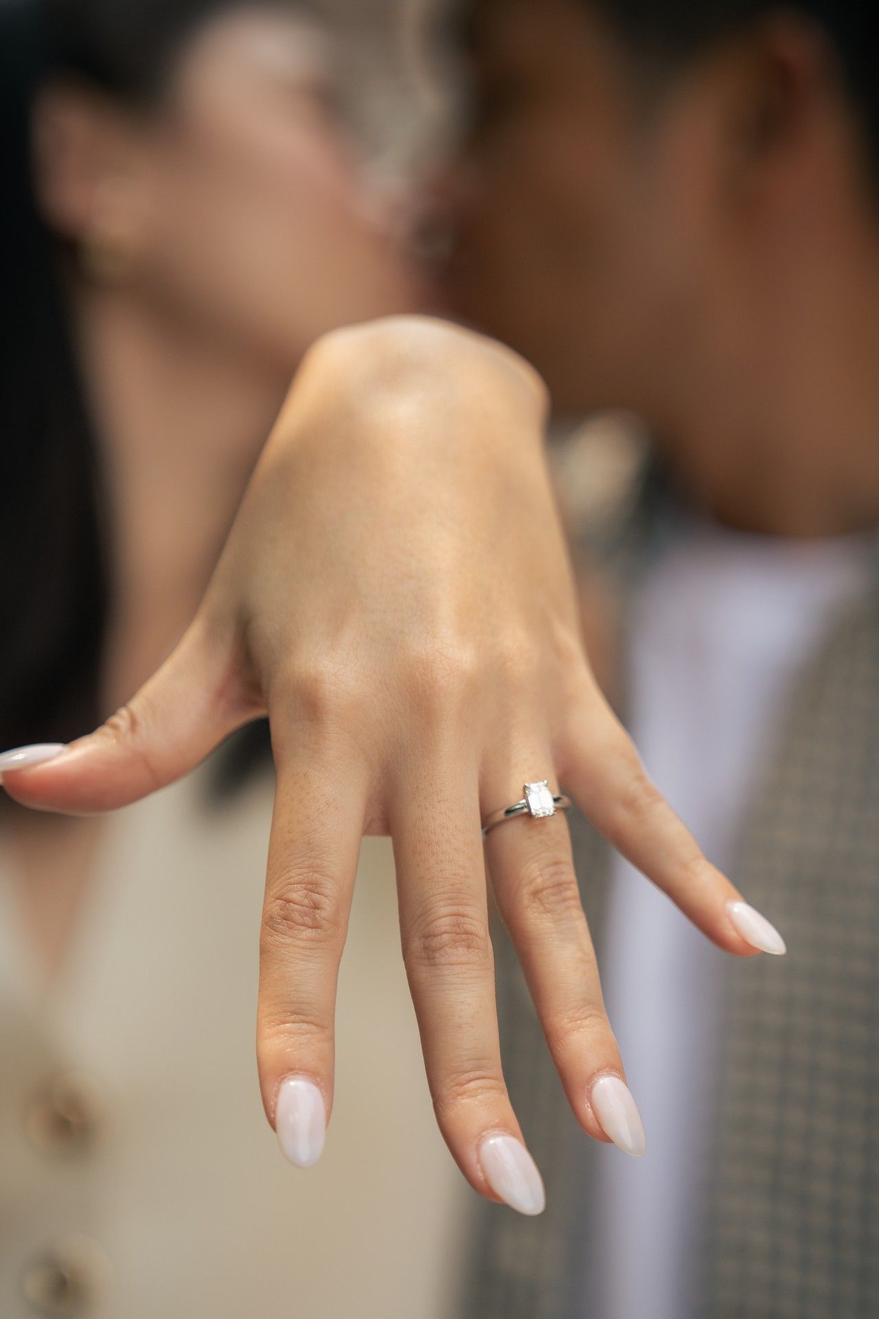 A hand wearing a white manicure displays a sparkling diamond engagement ring on the ring finger.
