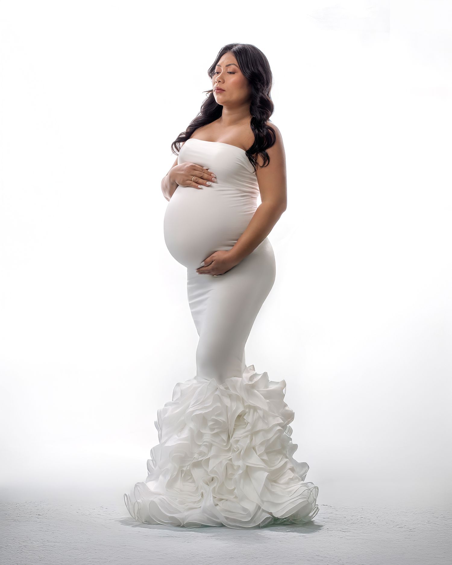 Beautiful pregnancy photo by Caz Captures of a woman in white modern gown looking serene against a white backlit backdrop.