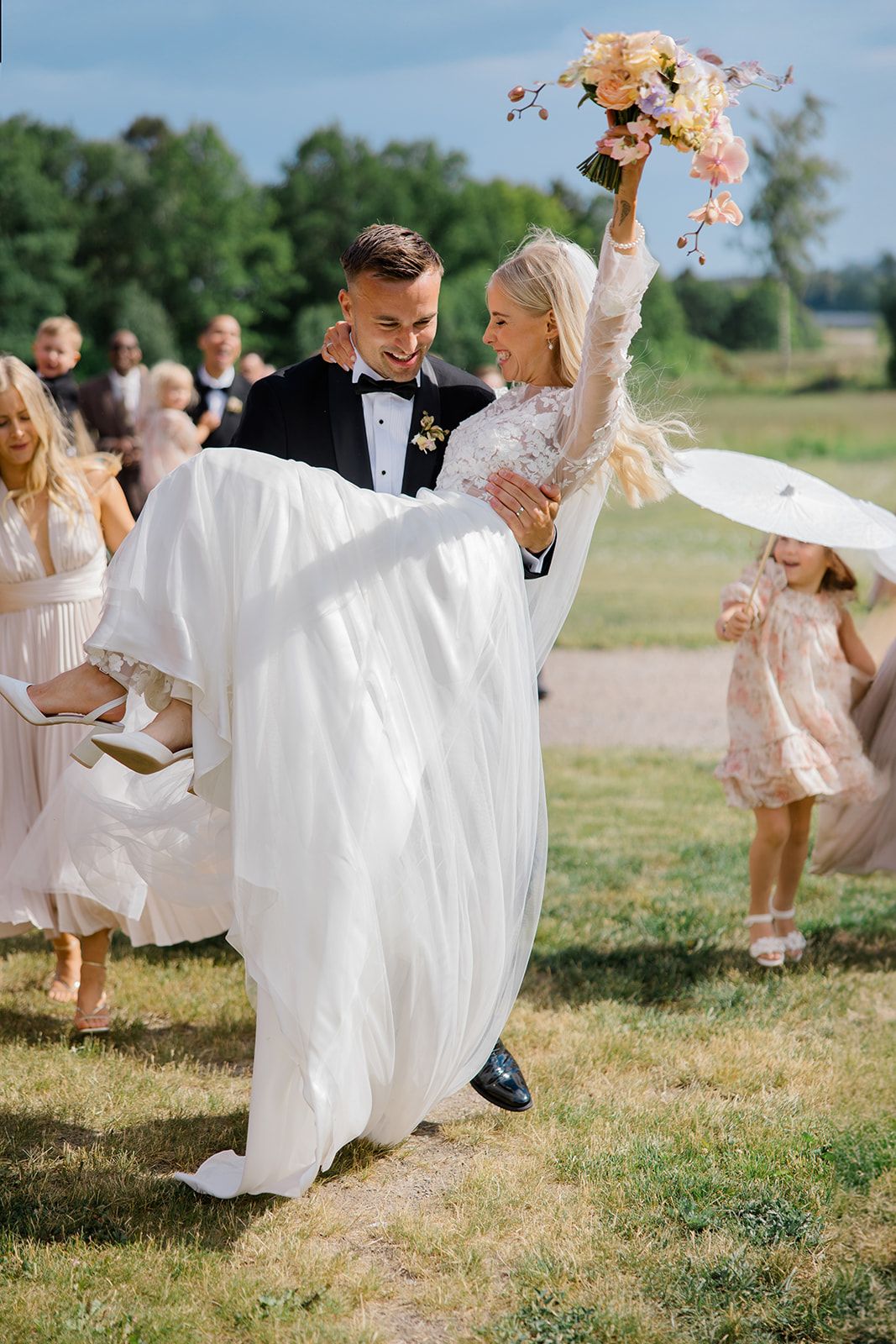 A joyful wedding party dances together on a sunny day in a grassy field, with flowing white dresses catching the breeze.