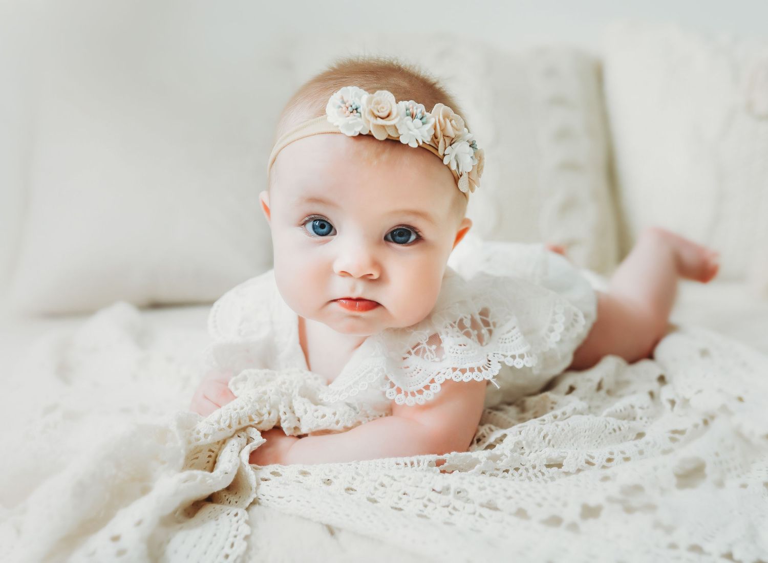A baby wearing a delicate white lace dress and floral headband poses on soft white bedding.