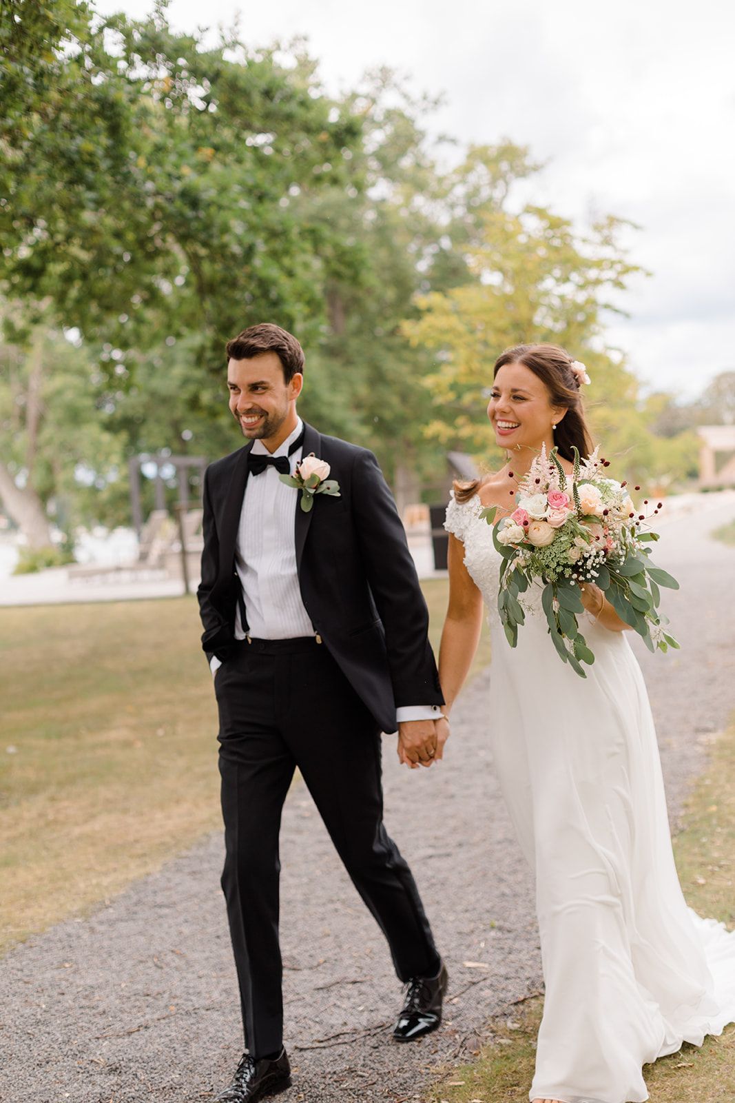 A joyful couple in wedding attire walks and laughs together on a gravel path surrounded by green trees.