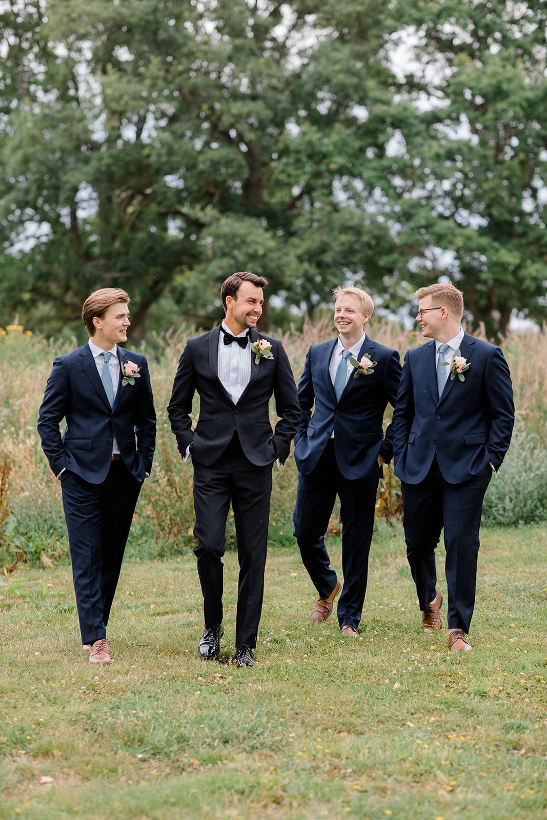 Groomsmen in navy suits laugh and walk together across a grassy field during wedding photos.