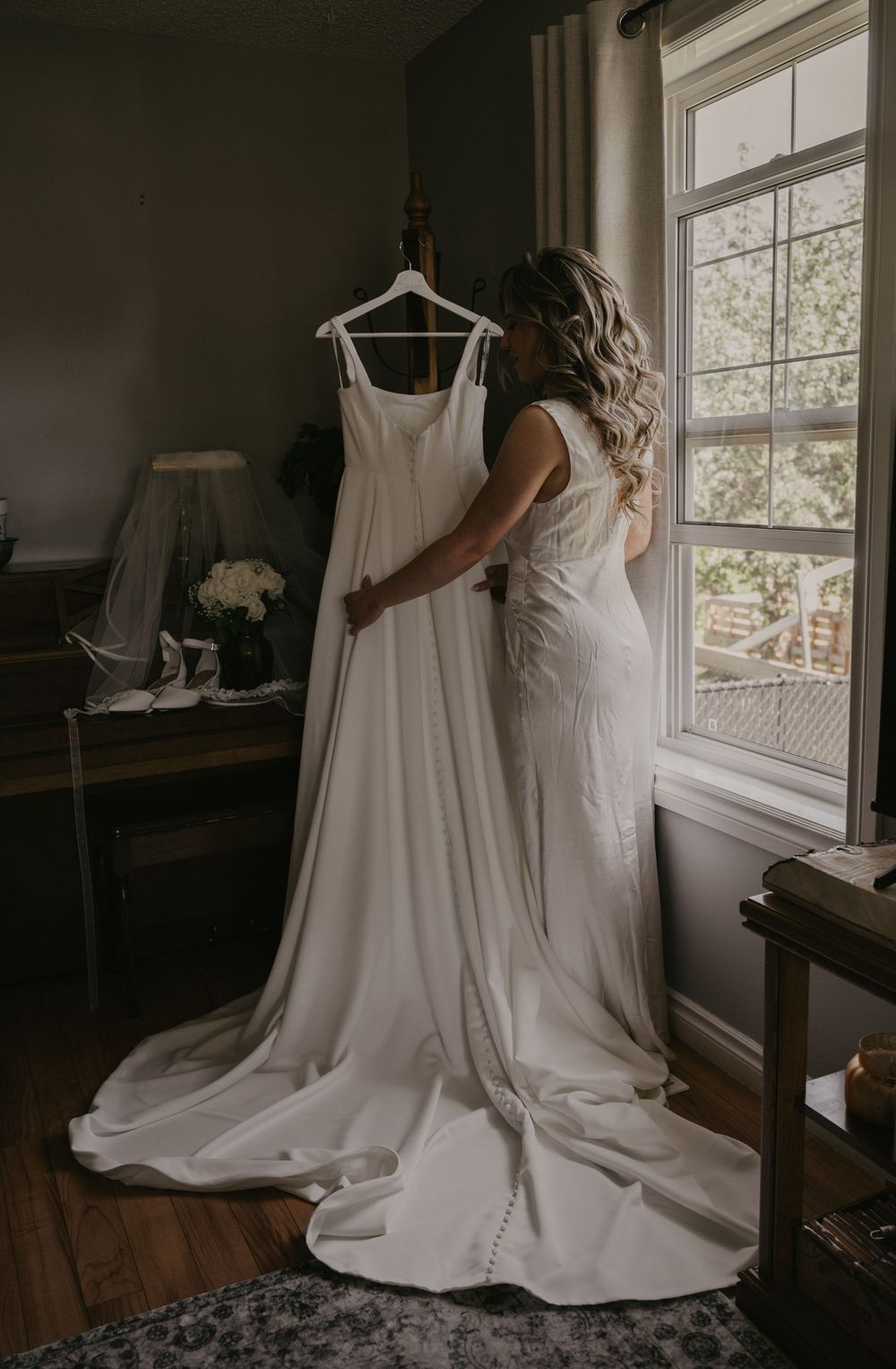 A wedding dress hangs by a window while someone in white admires it in a sunlit room.