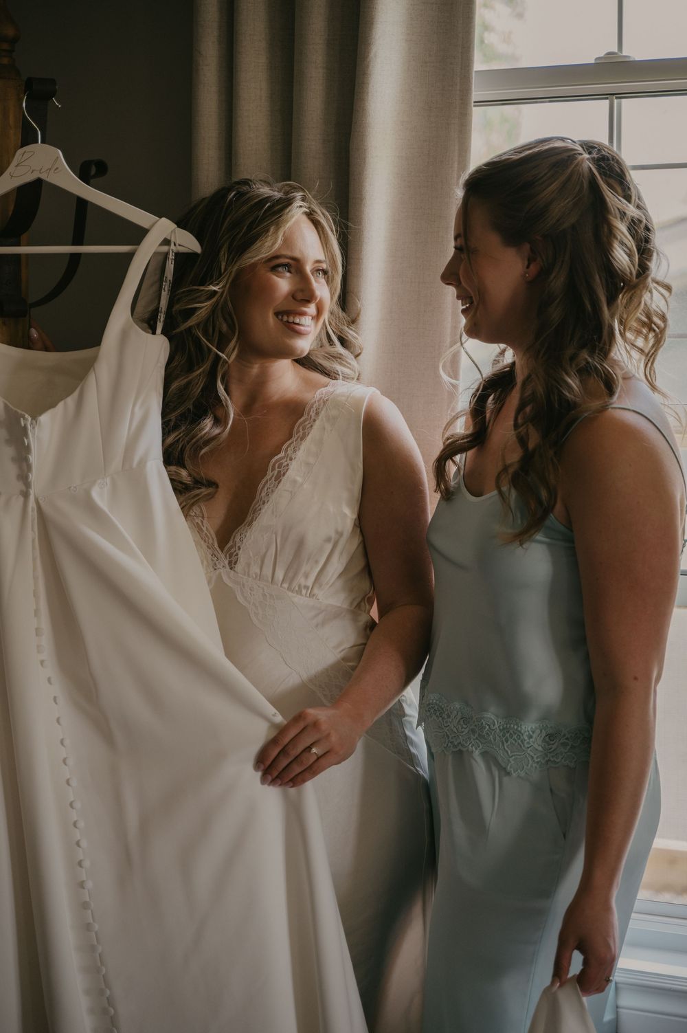 Bridesmaids in light blue dresses gather around a hanging white wedding gown by a window.