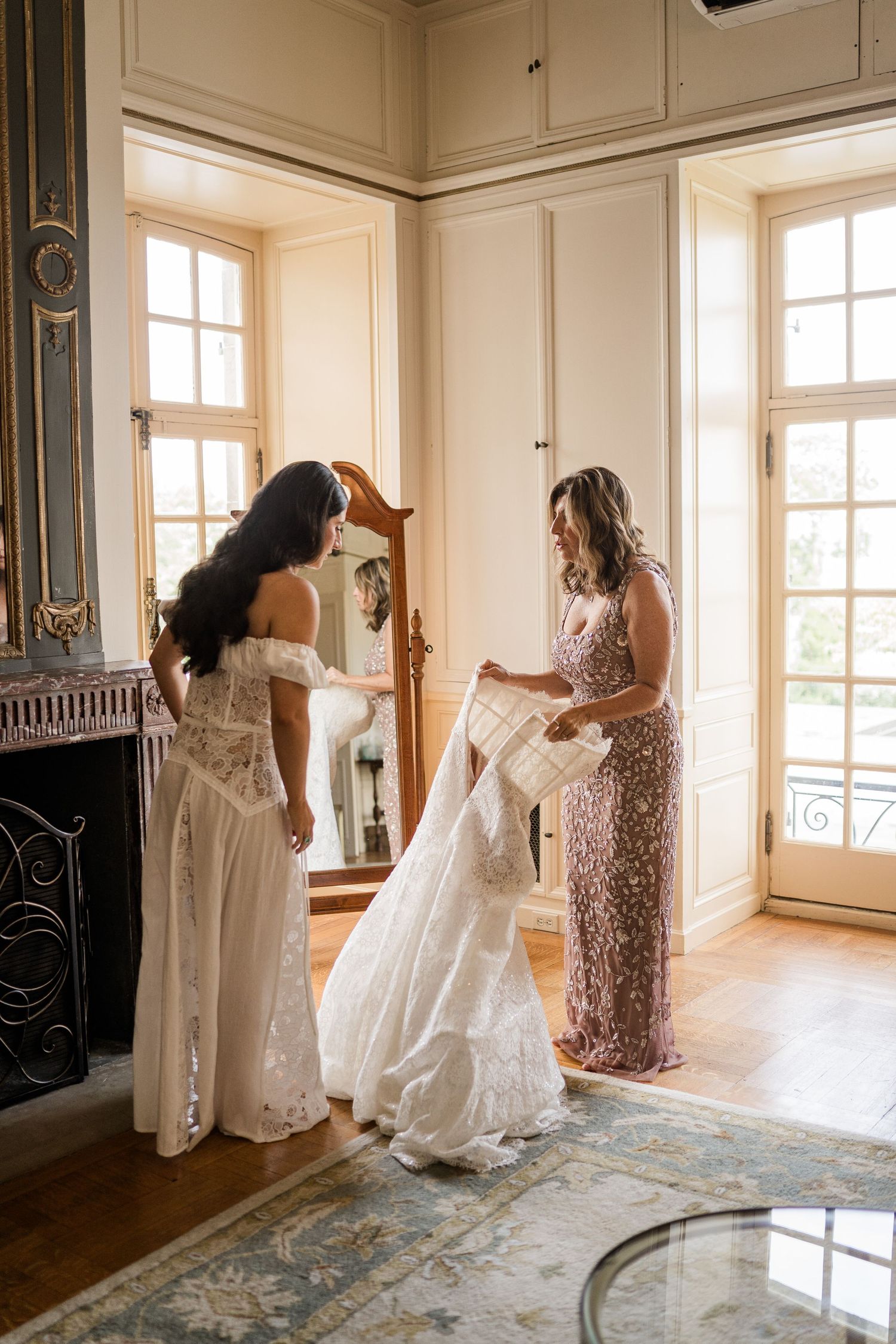 Two people prepare a wedding dress in a sunlit room with ornate details and vintage decor.