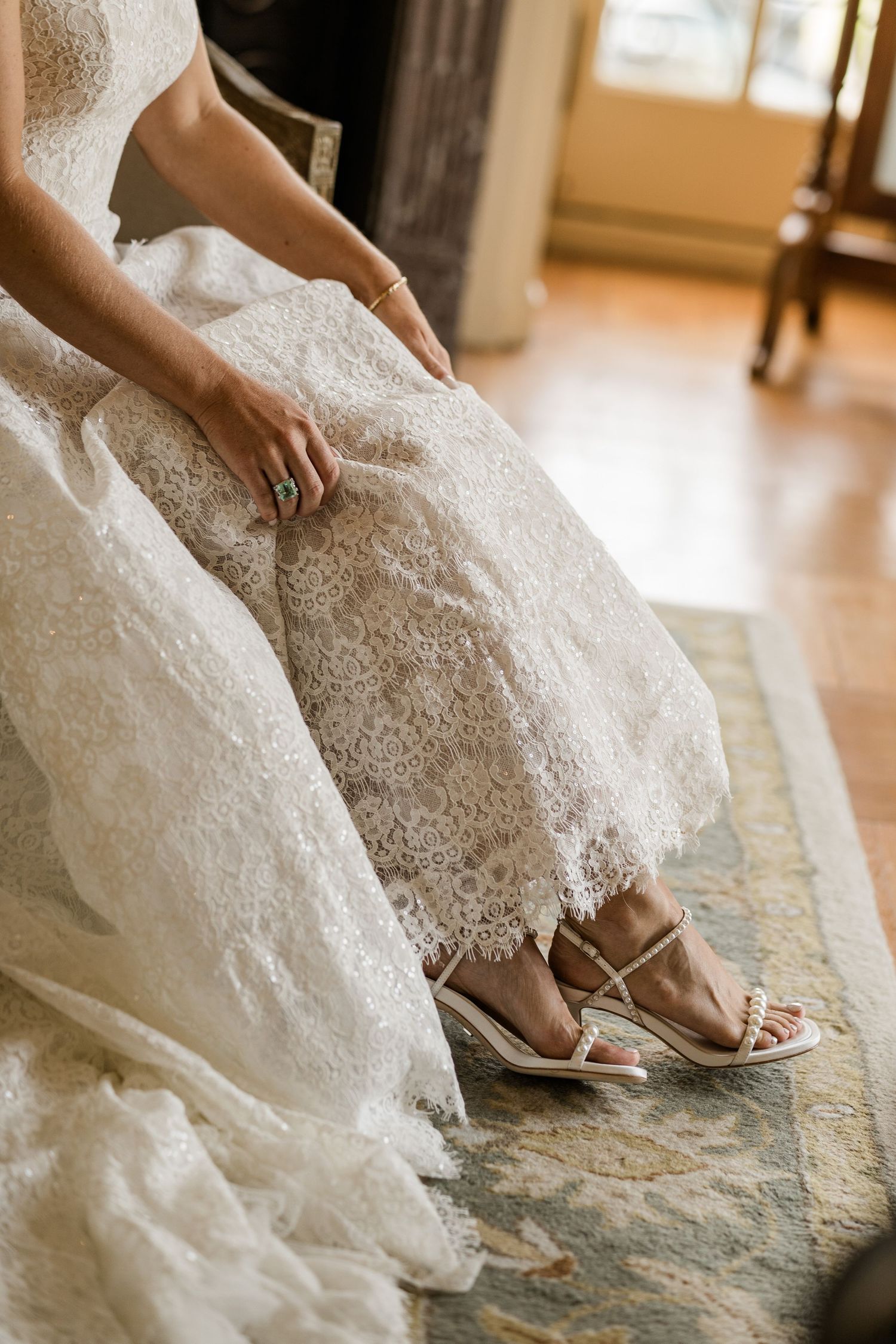 Close-up view of a bride's feet in silver sandals peeking out from under a white lace wedding dress.