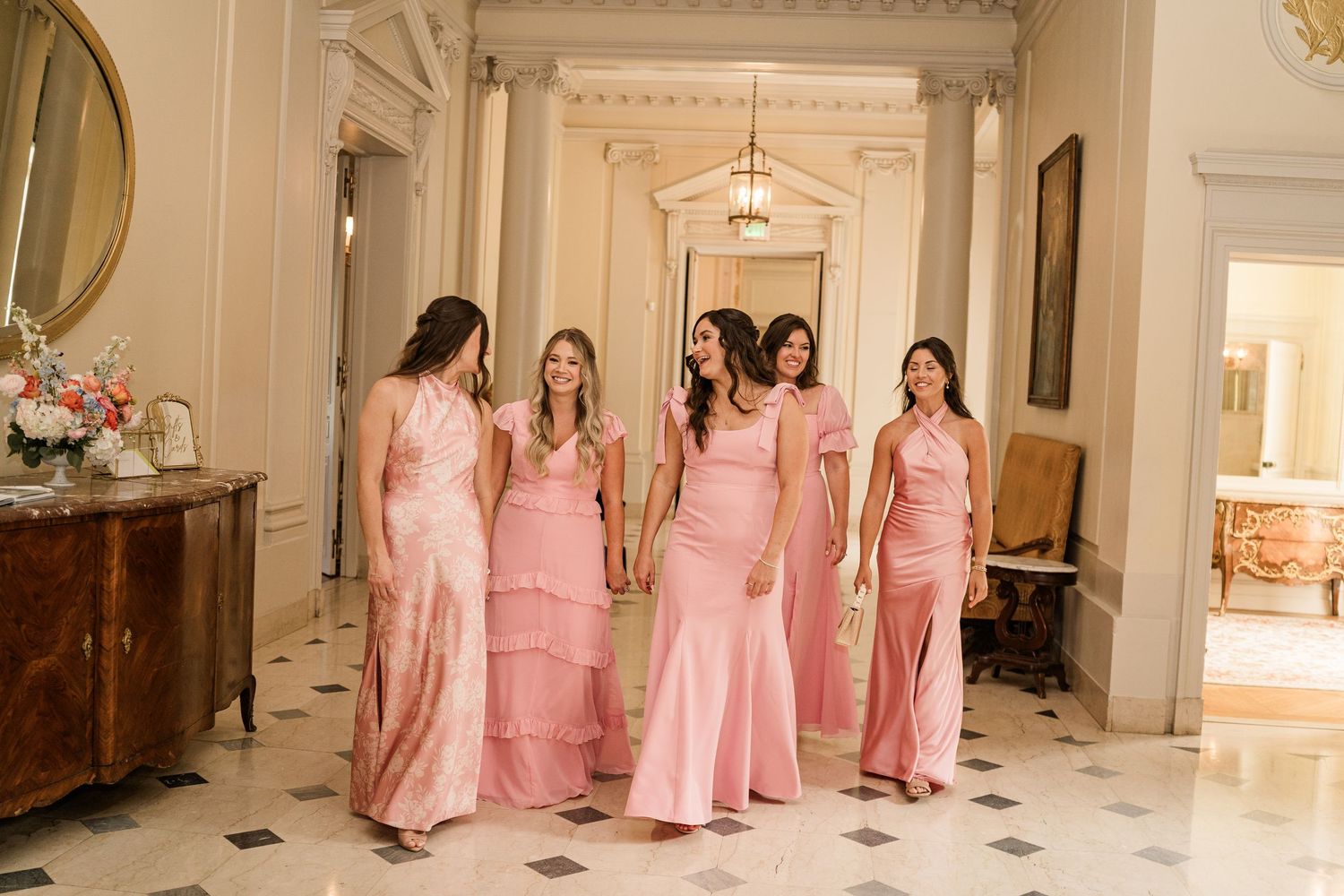 Bridesmaids in matching pink floor-length dresses walk together down an elegant hallway with marble floors and ornate decor.