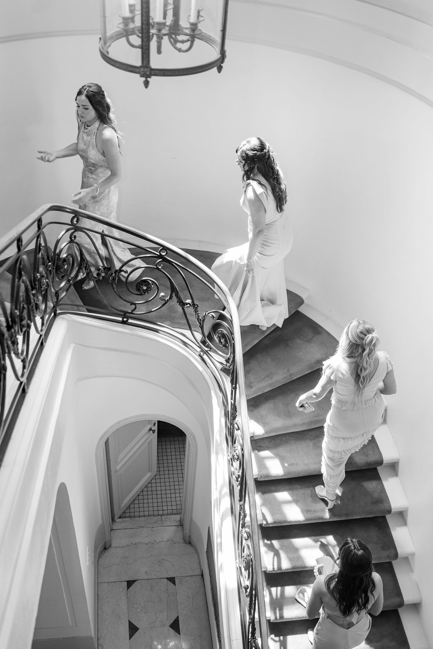 Four people in white outfits descend an elegant spiral staircase with ornate wrought iron railings under a hanging light fixture.