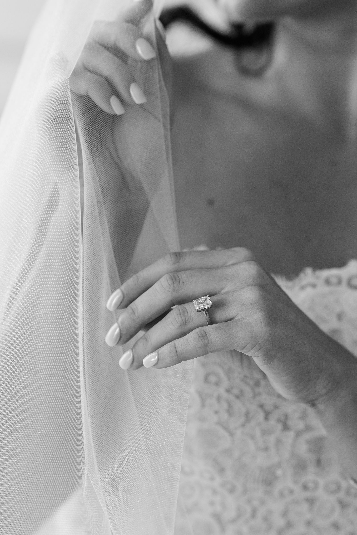 A close-up black and white photograph shows hands adjusting a delicate white lace veil with an engagement ring visible.