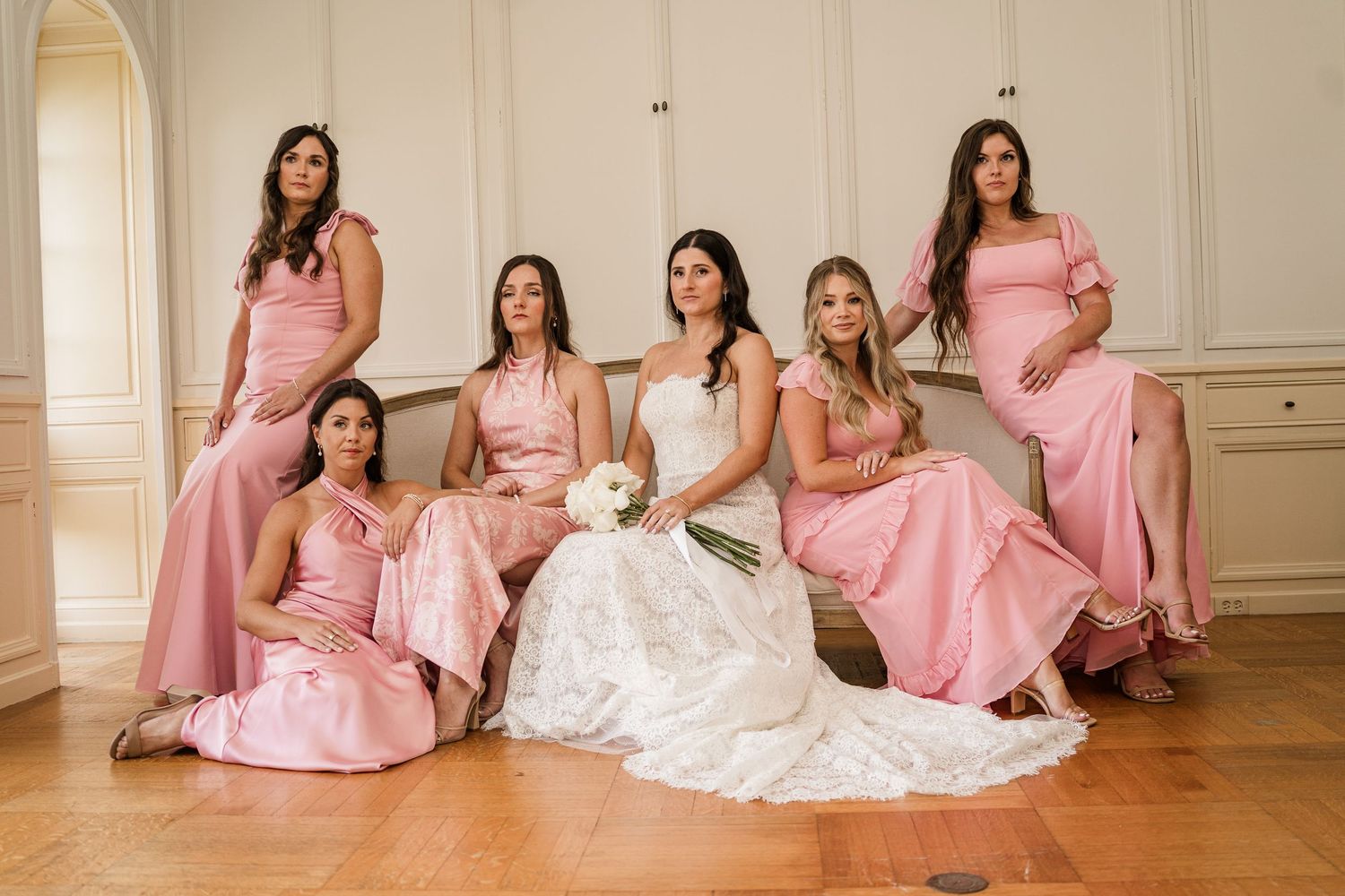 Wedding party in pink bridesmaid dresses poses together on hardwood floor in elegant room with white walls.