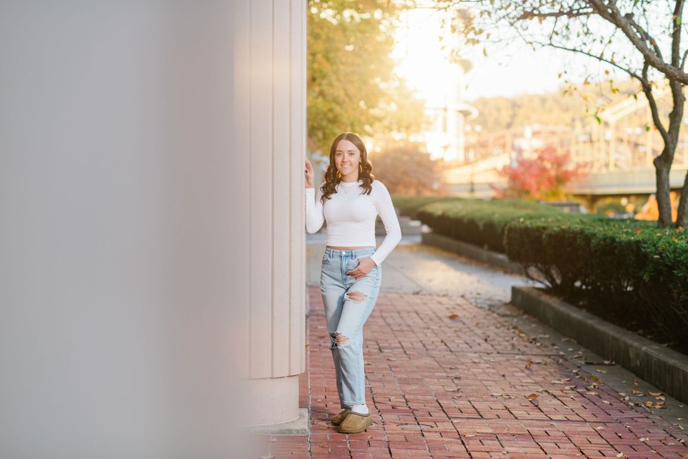 A person in a white sweater and light jeans stands against a wall on a brick path during golden sunset hours.