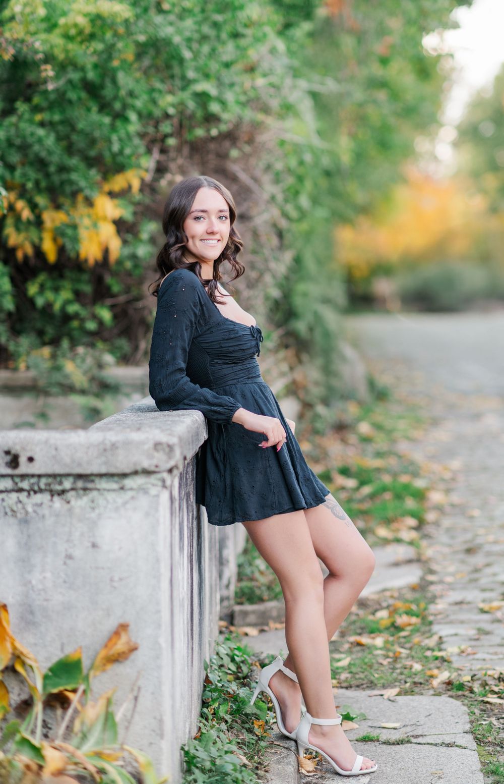 A figure in a black dress and silver heels poses along a stone wall surrounded by autumn foliage.