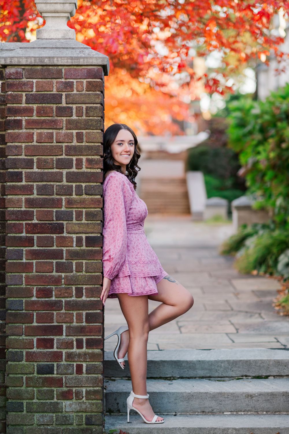 Woman in pink long sleeve dress poses on brick stairs with autumn foliage in background.