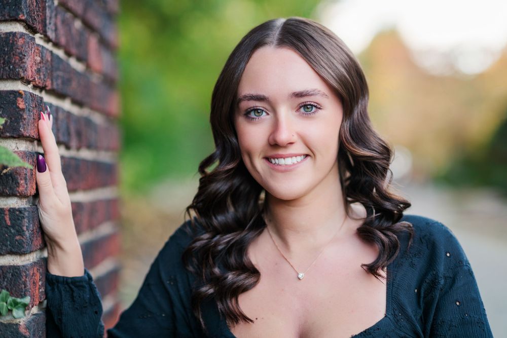 Young person in a navy cardigan poses next to a brick wall with soft natural bokeh in the background.