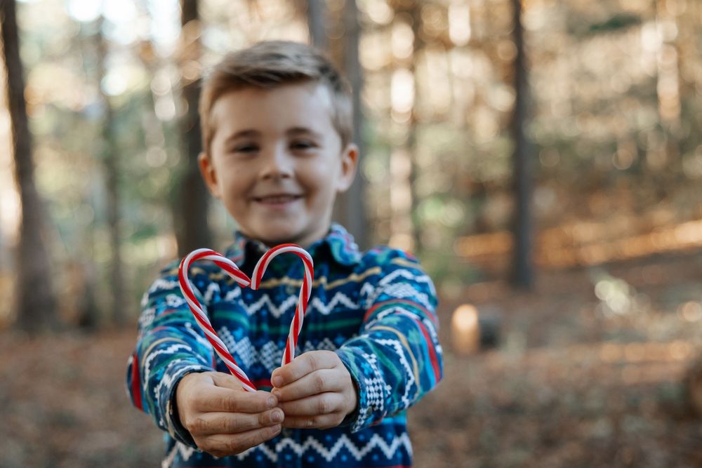 Child wearing blue patterned sweater holds candy canes in shape of heart against autumn forest background.
