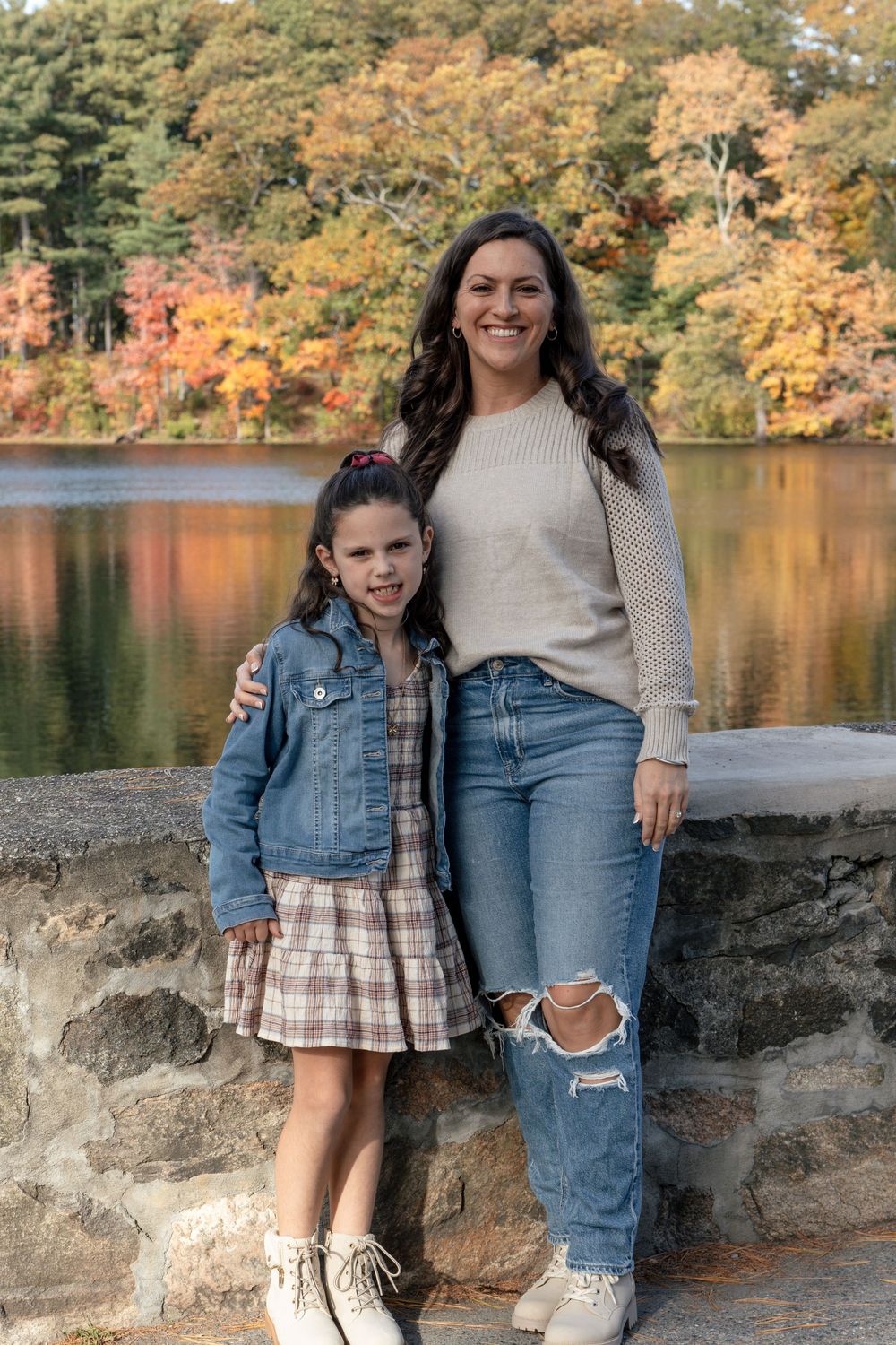 Mother and daughter in casual wear pose together on stone wall by autumn lake.