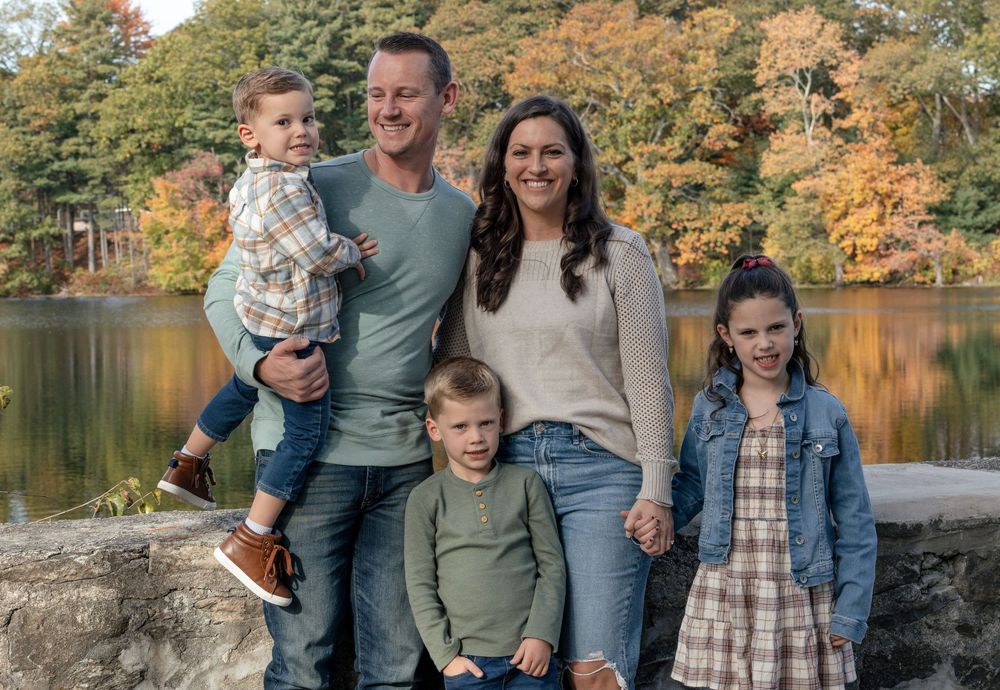 Family of five poses together on stone wall with autumn foliage and lake in background.