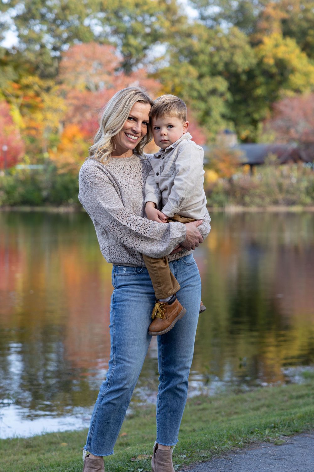 Mother in grey sweater holds child against autumn lake background.