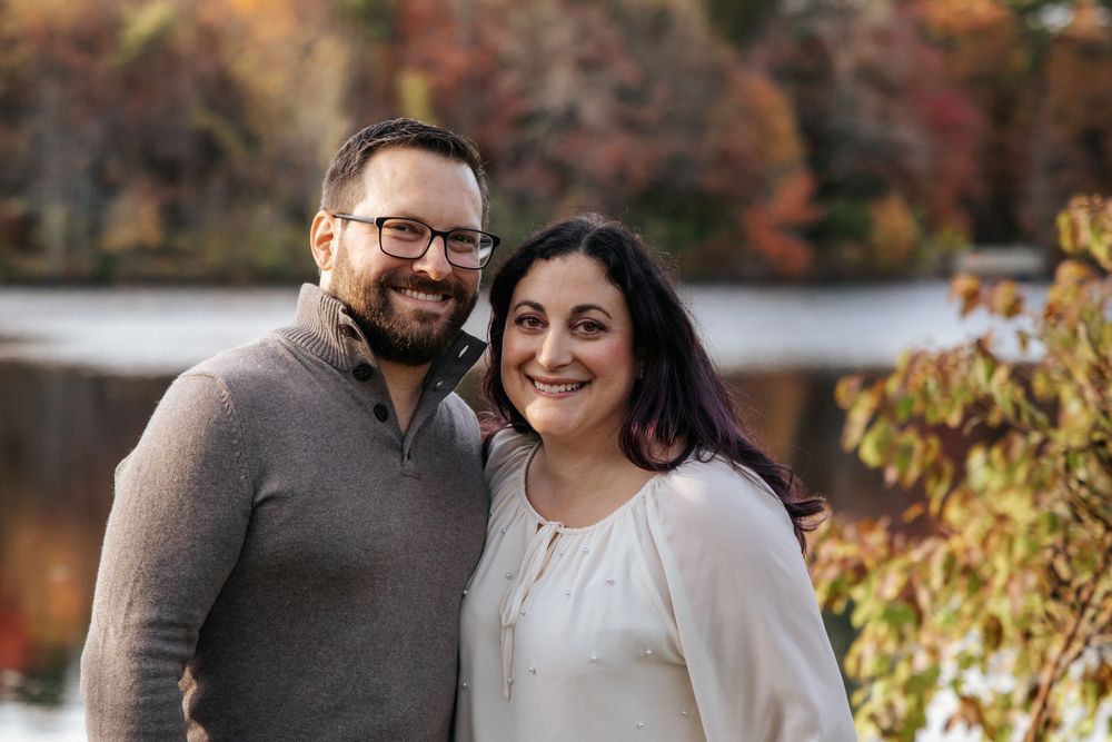 Couple in casual sweaters poses together with fall foliage and lake in background.