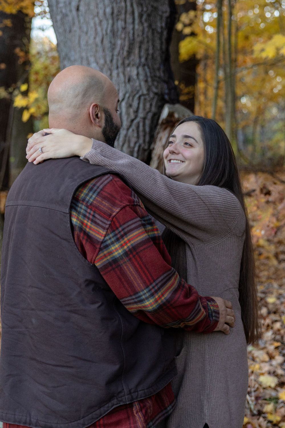 Couple embraces playfully in autumn forest setting with fallen leaves and trees in background.