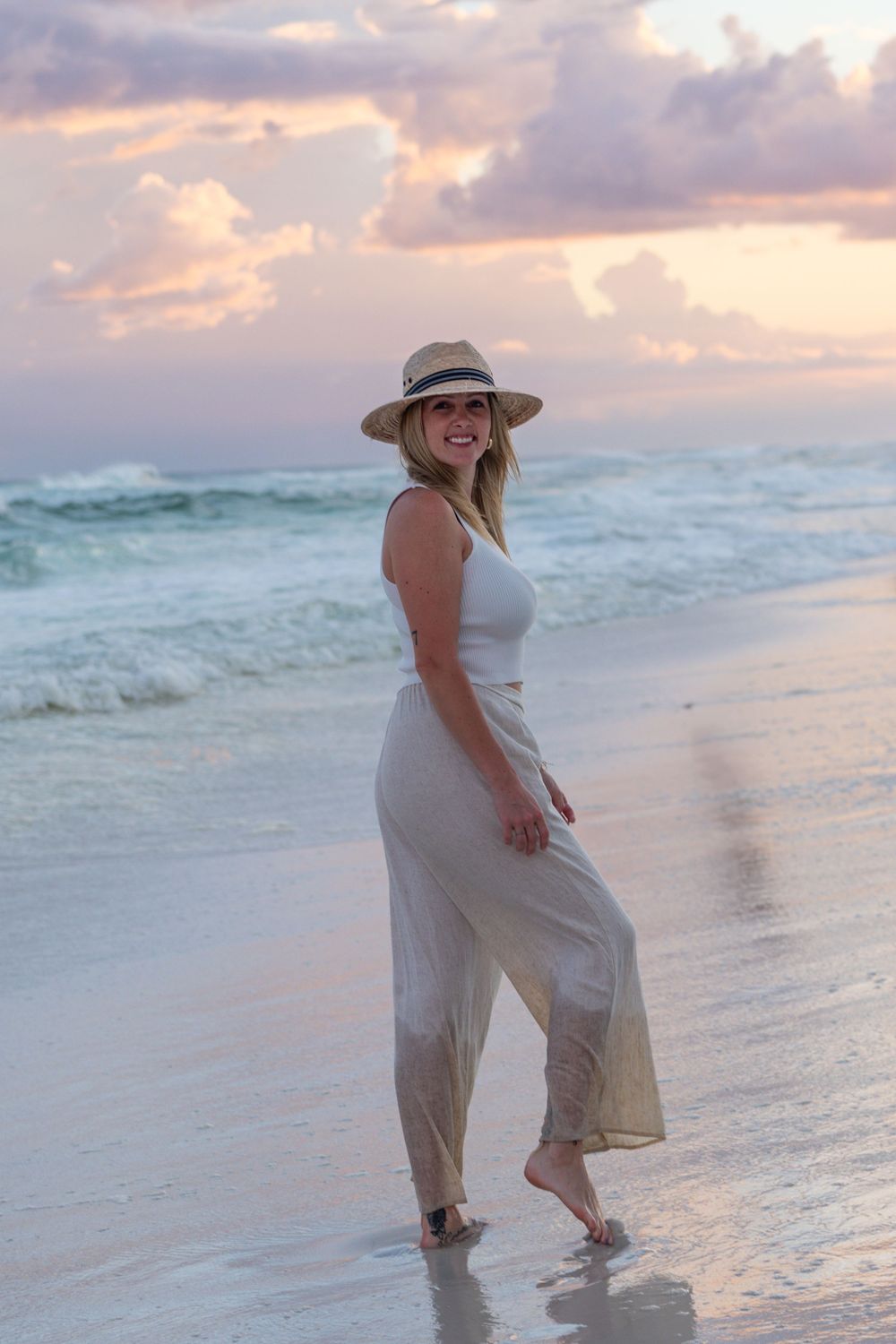 Woman in flowing dress walks along beach at sunset wearing wide-brim hat.