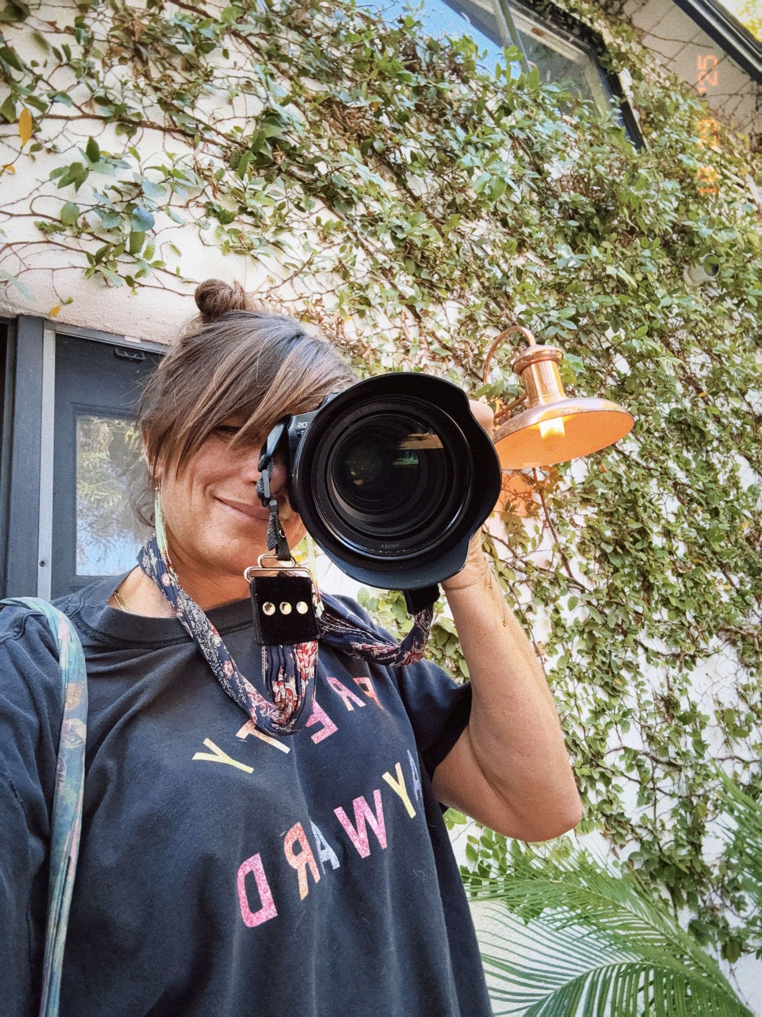 Photographer holds a professional DSLR camera with large lens outdoors against a leafy green backdrop.