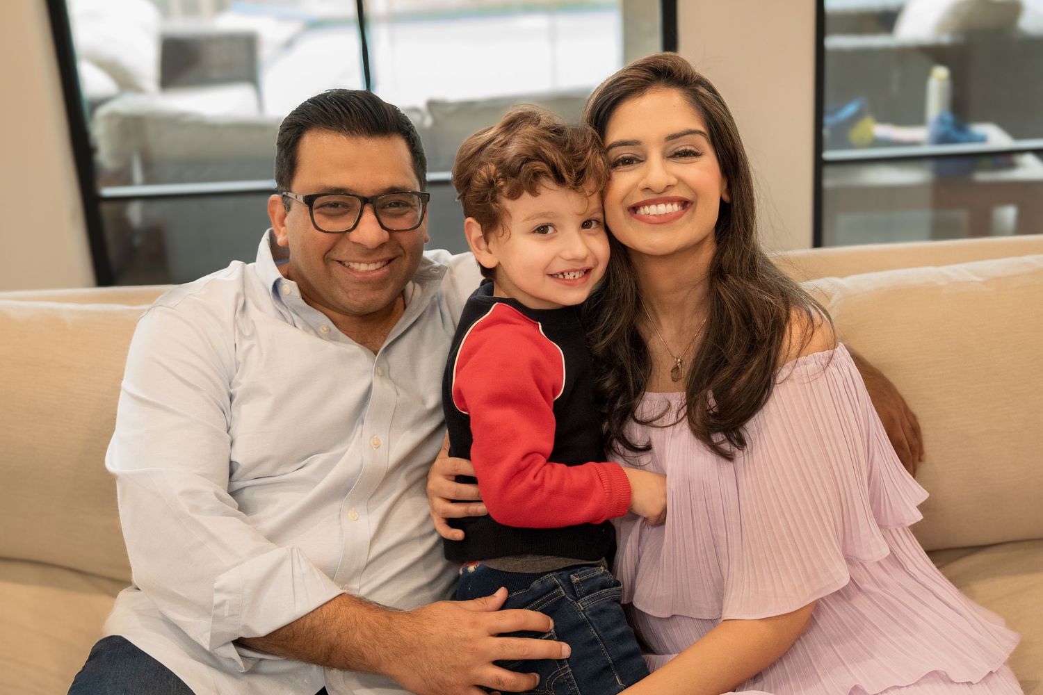 A family of three sits together smiling on a beige couch in a bright indoor setting.