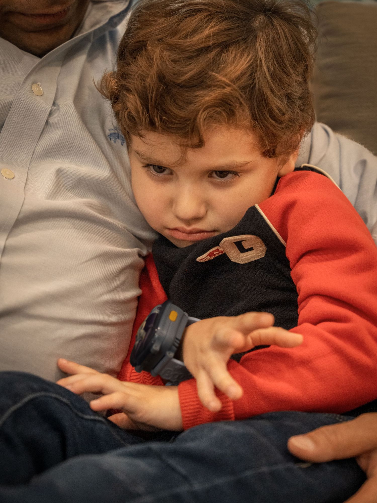 A child in a bright red sweater leans against someone while appearing to be in a sullen or grumpy mood.