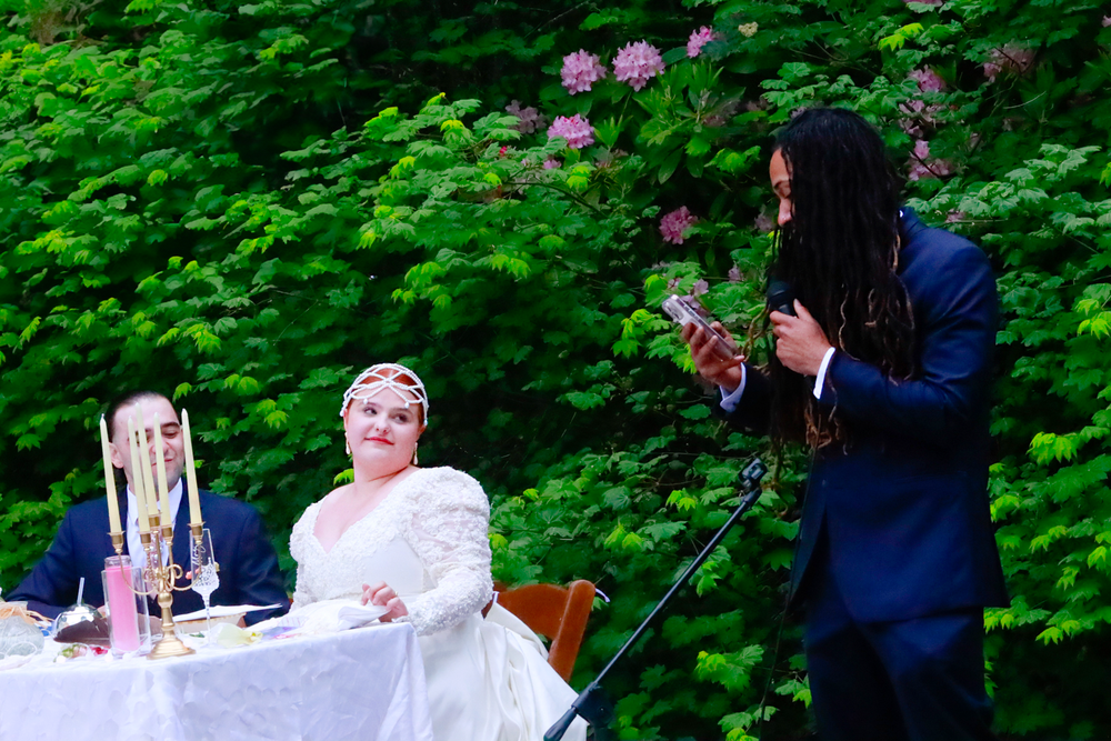 Guests mingle outdoors at a wedding reception surrounded by lush green foliage and pink flowers.