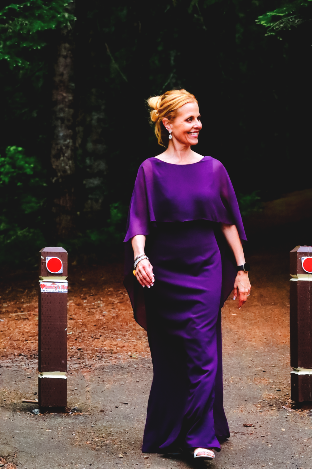 A person in a flowing purple dress poses near wooden posts in a forest setting.