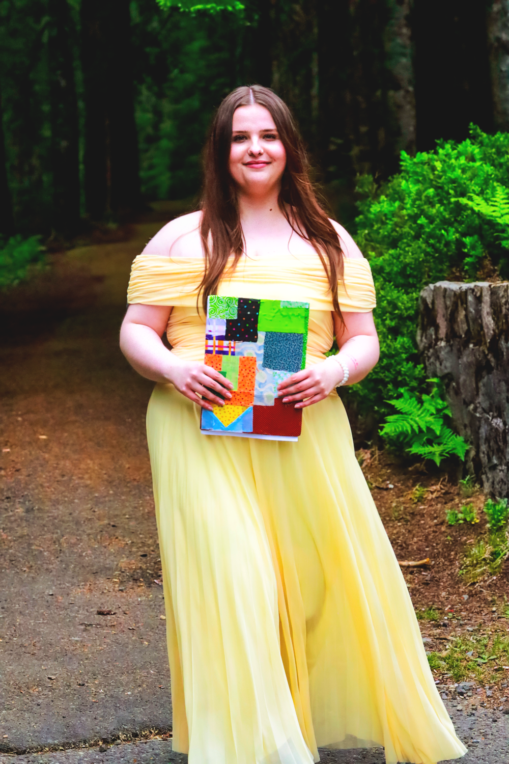 Person in yellow off-shoulder gown holding a colorful book while standing on forest path.