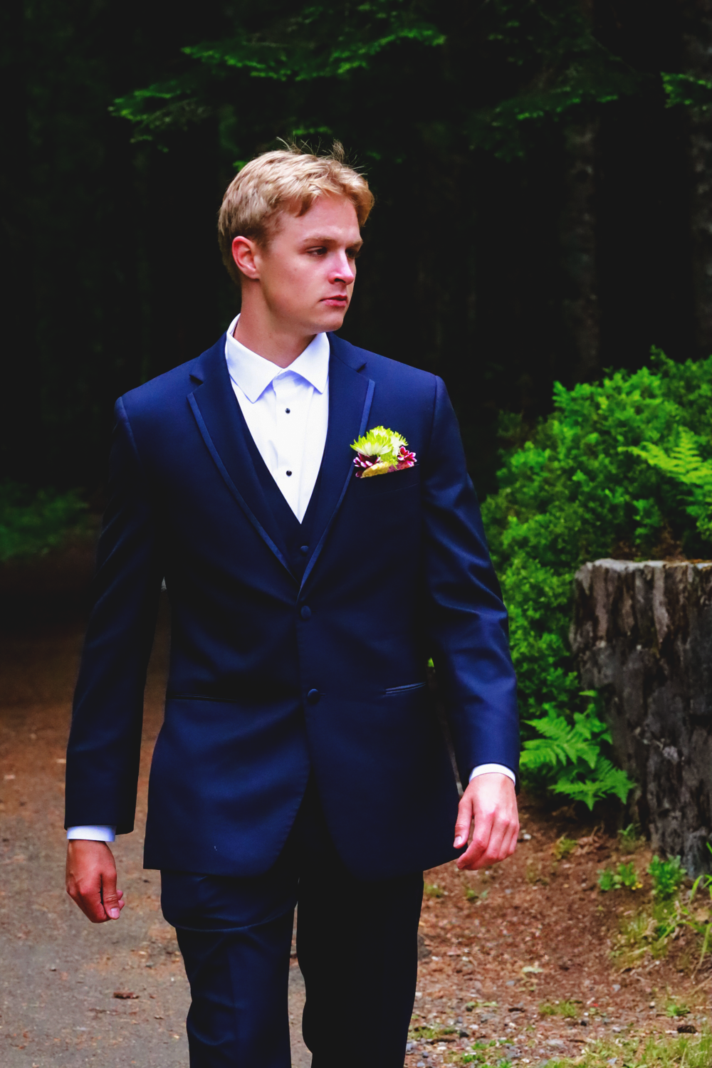 Person wearing navy blue tuxedo with yellow boutonniere standing on forest path.