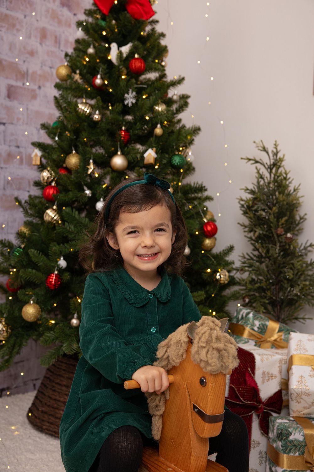 A festive Christmas scene with a decorated tree, presents, and a wooden rocking horse against a brick wall.