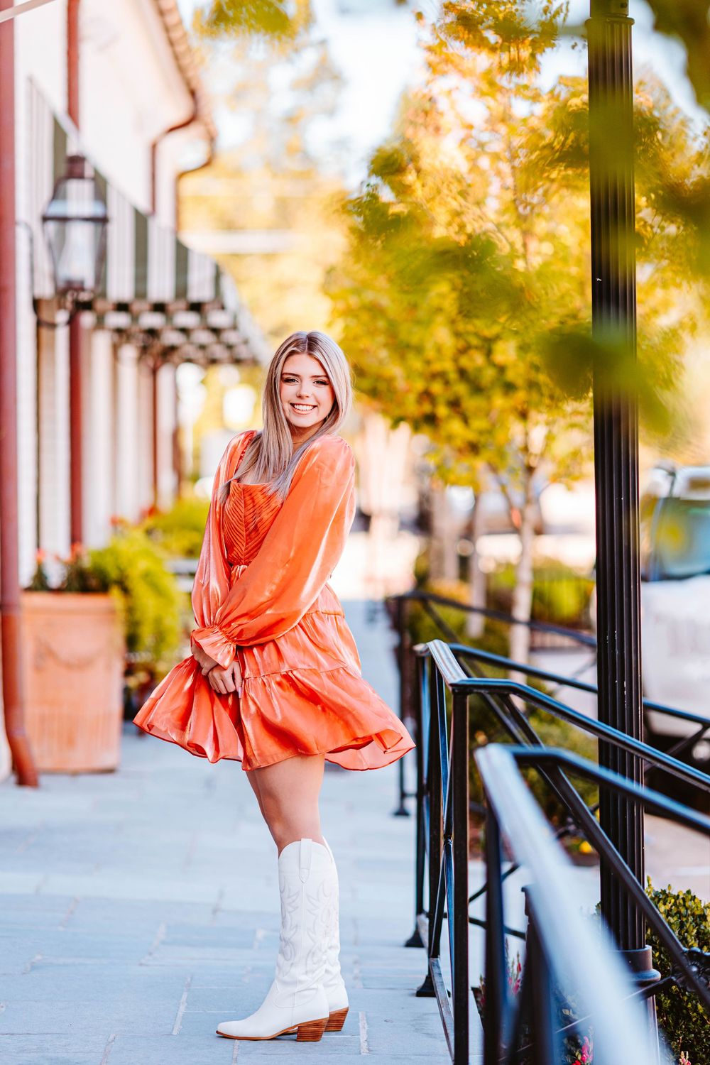 Person in coral dress and white boots poses playfully on urban sidewalk.