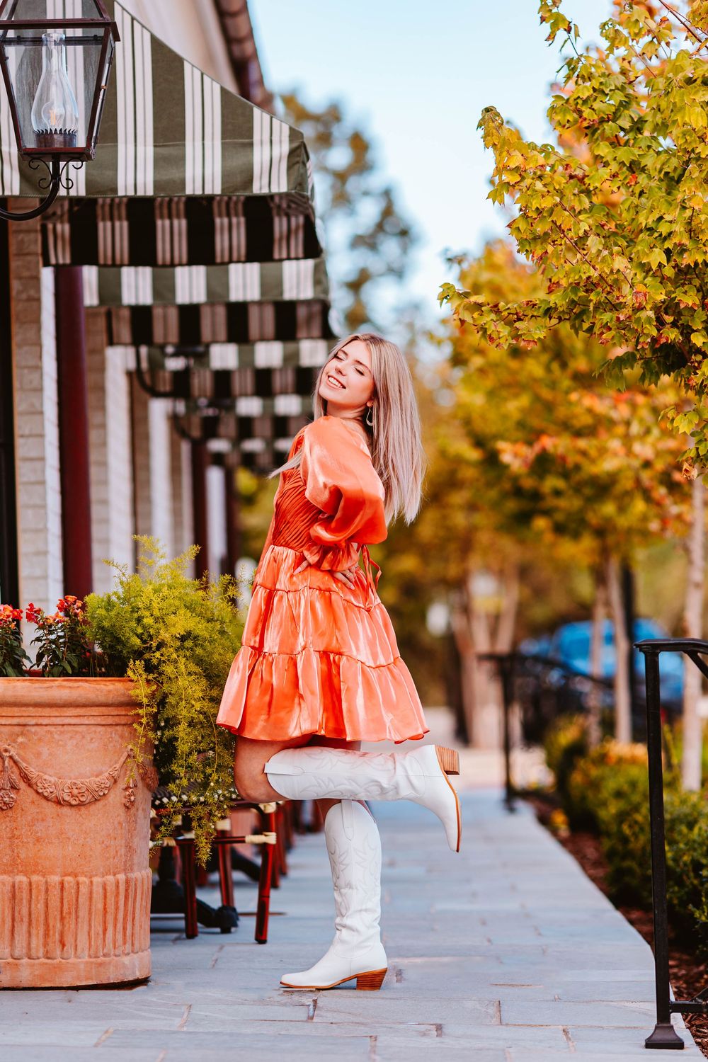 Person in bright orange dress dances on city sidewalk with autumn trees in background.