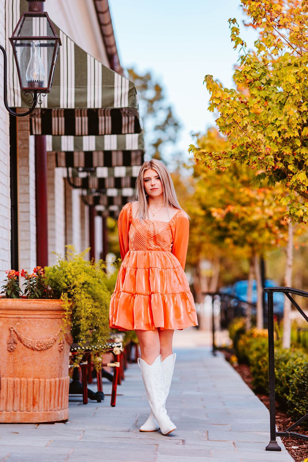 Person in ruffled orange dress poses against white building with autumn foliage.