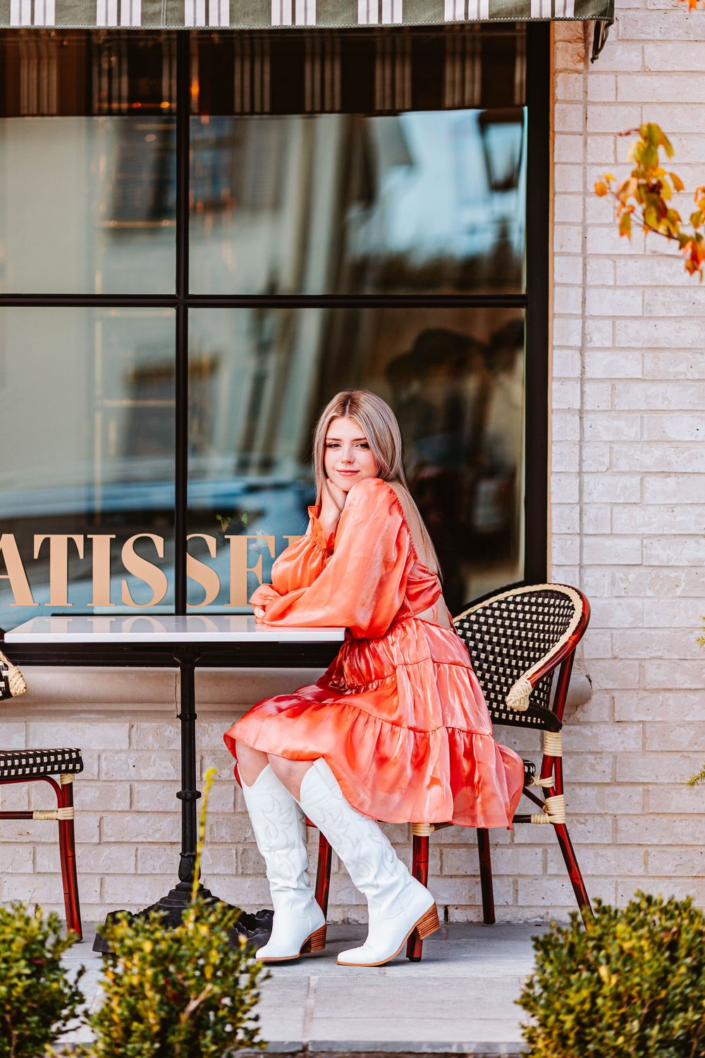 Person in orange dress and white boots poses on cafe patio.
