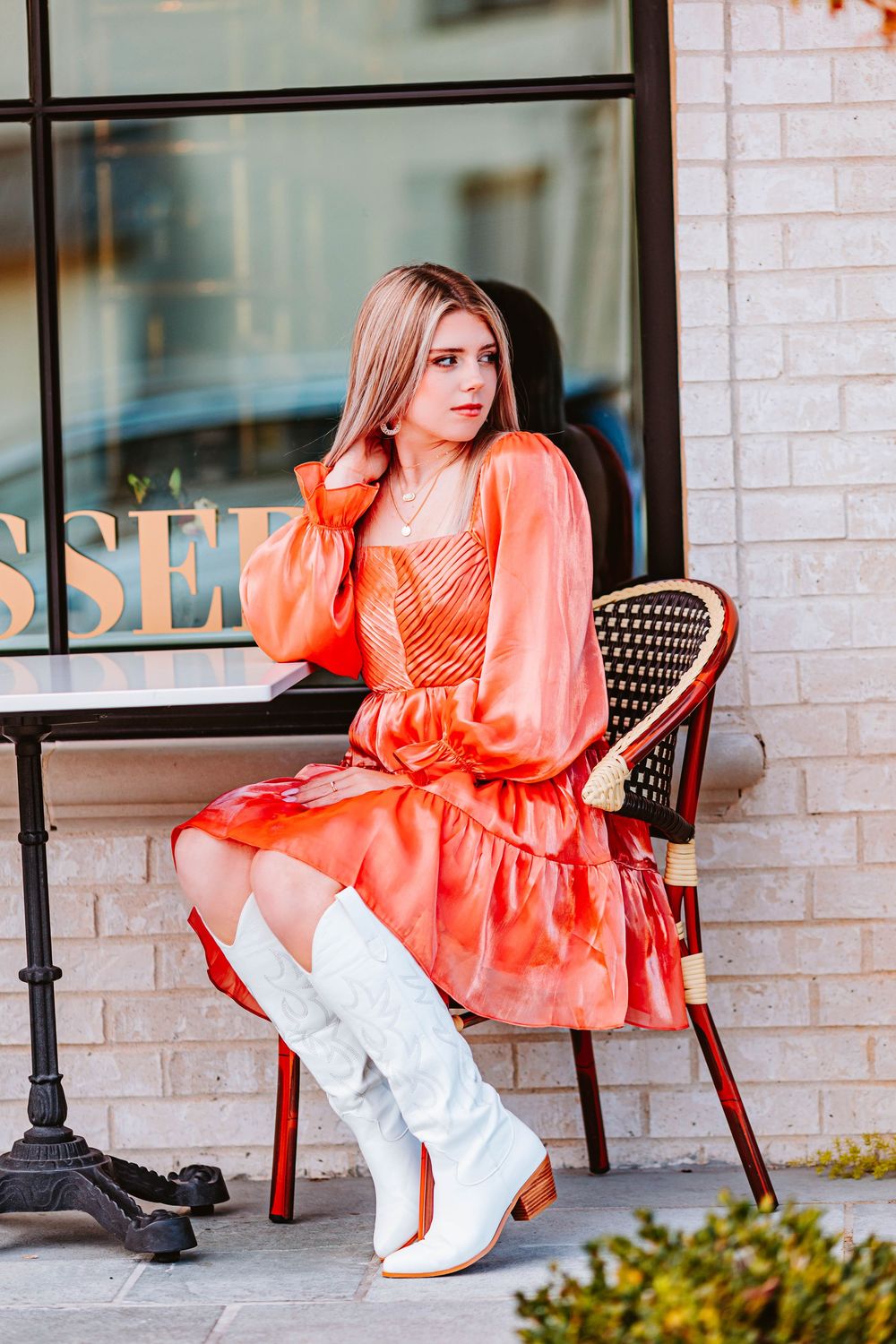 Fashion portrait of a person in a coral ruffled dress and white boots sitting on outdoor cafe chairs.